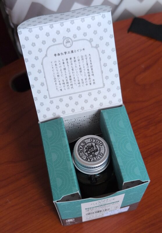 Open box of Teranishi Guitar fountain pen ink, showing the nice box design where cardboard padding holds the top of the ink bottle safely padded for shipping. The lid of hte bottle has a sticker on it that reads "guitar gold star" and has their music note logo with other shape in the center.