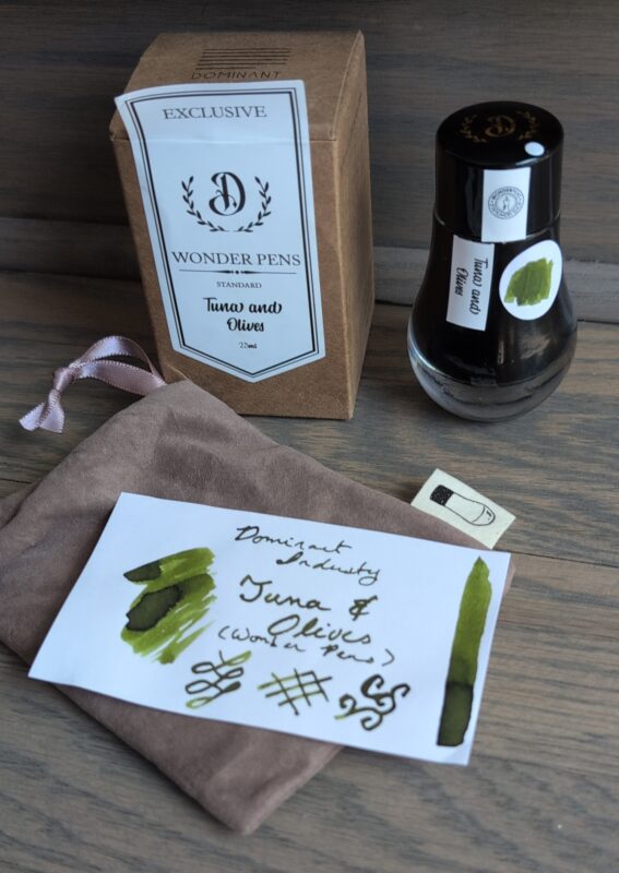 Dominant Industry Tuna and Olives Ink swatch and bottle with packaging. The bottle has a rounded base that makes it look like a water droplet, there's a bag with a picture of the bottle on the label, and a simple cardboard box. I've added a dot with an ink swatch to the side of the bottle, showcasing the olive green colour.