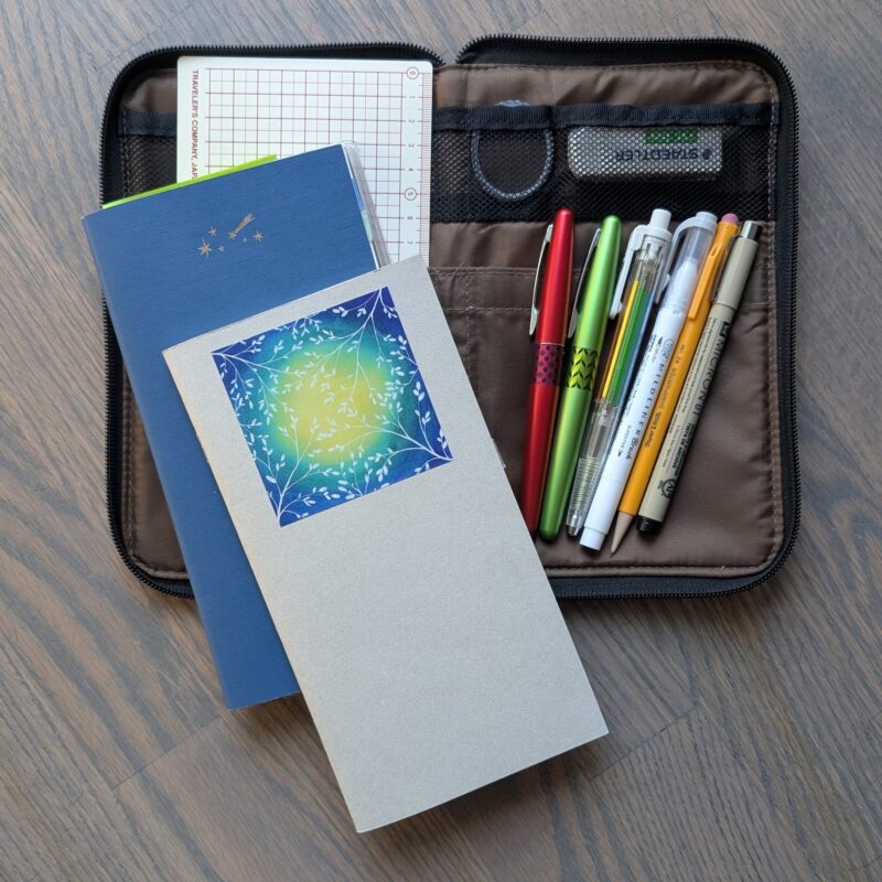 Traveler's notebook (dot grid, weekly planner, and blank), a grid pencil board, two fountain pens, a grey double-ended brush pen, a multi-colour pencil, a regular mechanical pencil, a black fineliner, an eraser, and a tiny roll of thin washi tape, all sitting on top of a large flat pencil case.