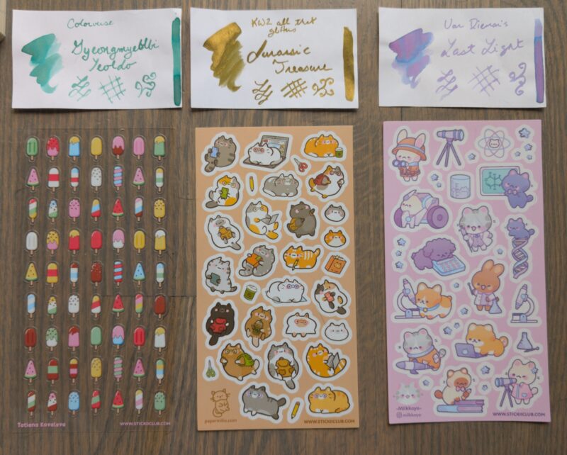 Inks and Stickers close up: Colorverse Gyeongnyeolbi Yeoldo (pale green), KWS all that glitters Jurrasic Treasure (glittery gold with very mildly green undertone), Van Dieman's Last Light (purple with blue shading), and sticker sheets with popsicles, school cats, and science animals.